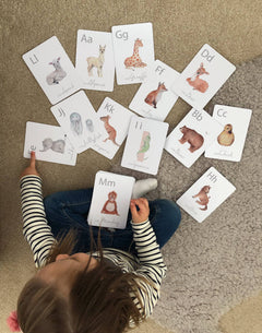 A child is playing with a set of white animal-themed alphabet flashcards with a letter on each card, featuring illustrations of various animals such as a giraffe, a zebra, a deer, and a bear.