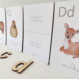 A child is playing with a set of white animal-themed alphabet flashcards with a letter on each card, featuring illustrations of various animals such as a giraffe, a zebra, a deer, and a bear.