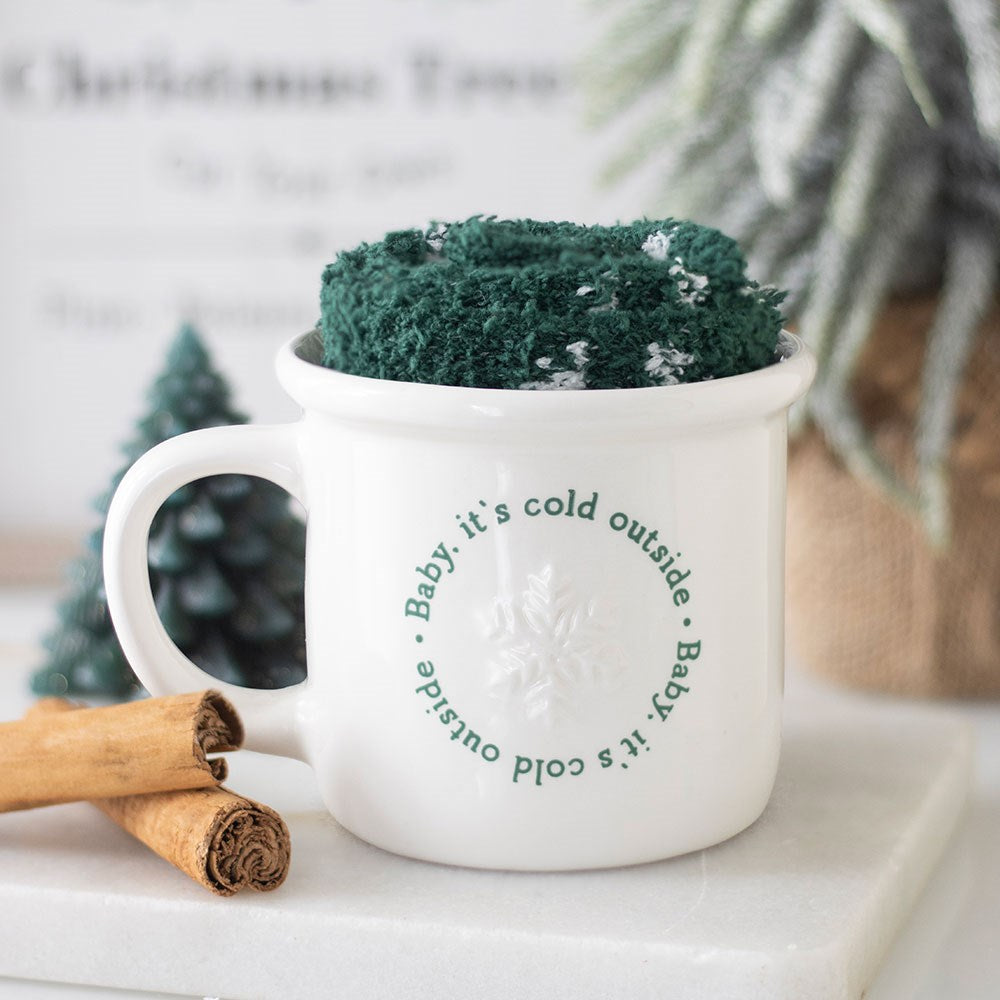 White mug with text and green knitted cozy on a white surface with cinnamon sticks and a small Christmas tree.