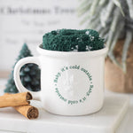 White mug with text and green knitted cozy on a white surface with cinnamon sticks and a small Christmas tree.