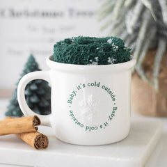 White mug with text and green knitted cozy on a white surface with cinnamon sticks and a small Christmas tree.