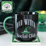 Black mug with 'Bad Witch Social Club' text and green witch hat design on a coaster, with 'Bad Witch Social Club' logo in top left corner.