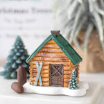 Miniature log cabin with green roof, skis, and small trees on a white surface.