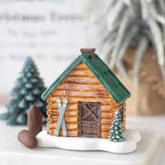 Miniature log cabin with green roof, skis, and small trees on a white surface.