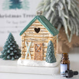 Decorative log cabin with a heart-shaped window, surrounded by small trees and an essential oil bottle.