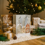 Decorative Christmas Gift Bag with a village scene in front of a Christmas tree and presents.