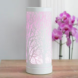 An electric wax melt warmer with a white tree silhouette design, placed on a table with a plant and a decorative oil lamp in the background, with text 'LED Colour Changing'.