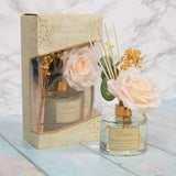 A floral diffuser bottle with a gold-colored cap, surrounded by a bouquet of magnolia and mulberry flowers, placed in front of its packaging box on a marble surface.