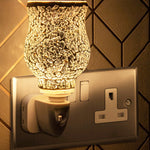 A silver mosaic plug-in warmer with a UK plug, mounted on a wall, emitting a glow.