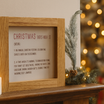 Framed Christmas definition sign on a mantelpiece with candles and a decorated tree in the background.