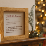 Framed Christmas definition sign on a mantelpiece with candles and a decorated tree in the background.