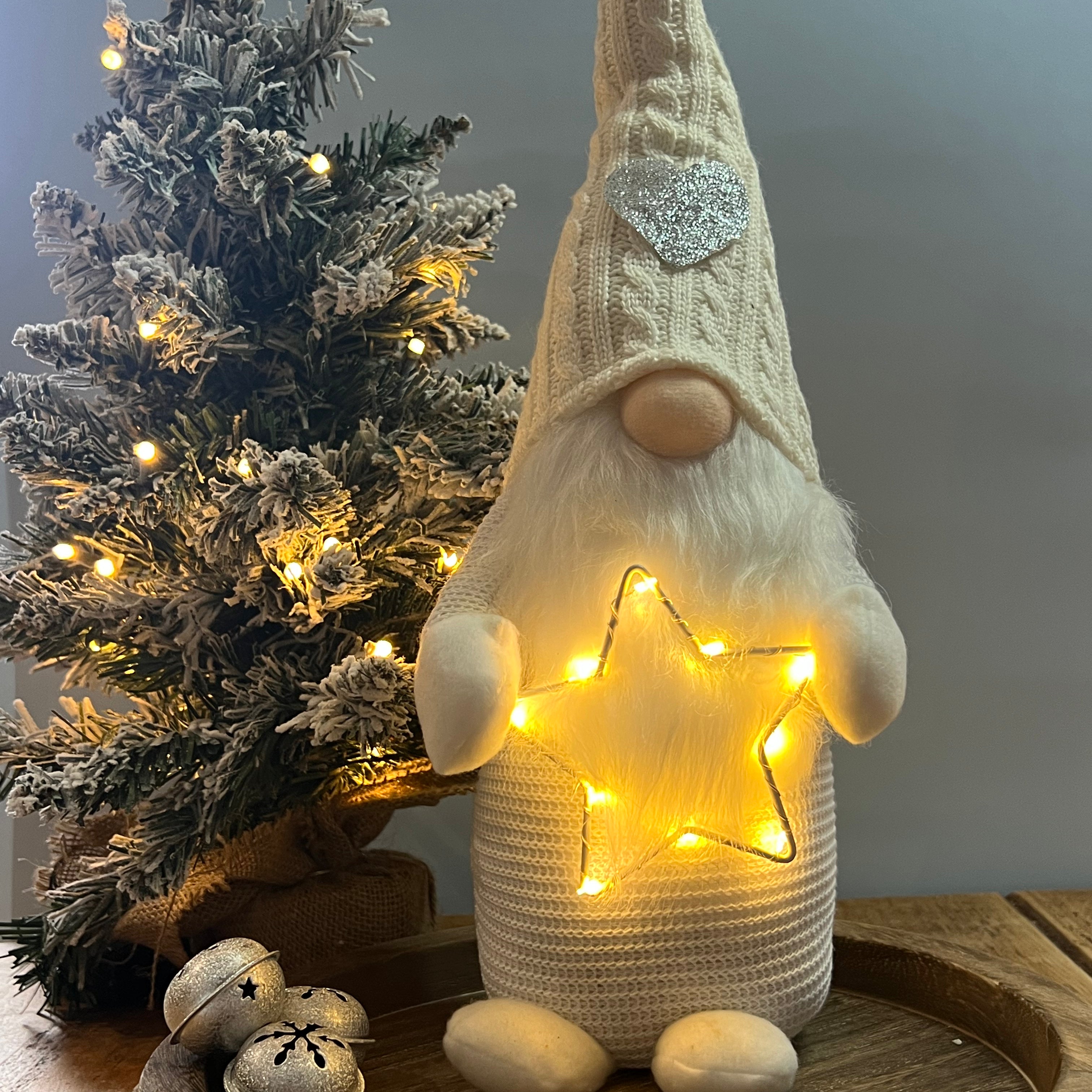 A standing decorative Gonk figure with a knitted hat and a LED star on its head, displaying a warm glow, placed next to a decorated Christmas tree and a small gift-wrapped present.