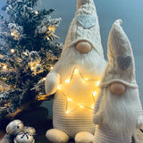 A standing decorative Gonk figure with a knitted hat and a LED star on its head, displaying a warm glow, placed next to a decorated Christmas tree and a small gift-wrapped present.