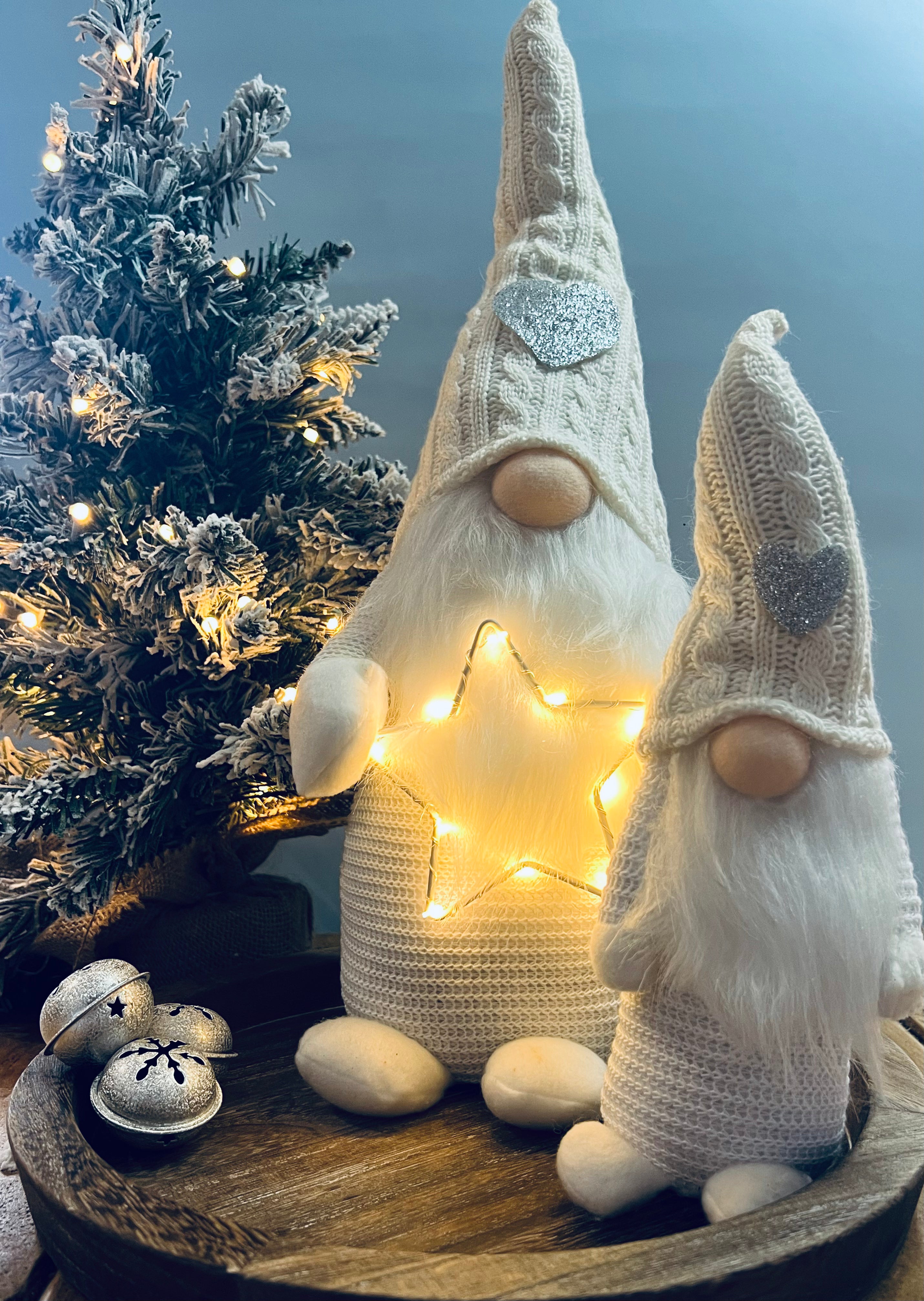 A standing decorative Gonk figure with a knitted hat and a LED star on its head, displaying a warm glow, placed next to a decorated Christmas tree and a small gift-wrapped present.