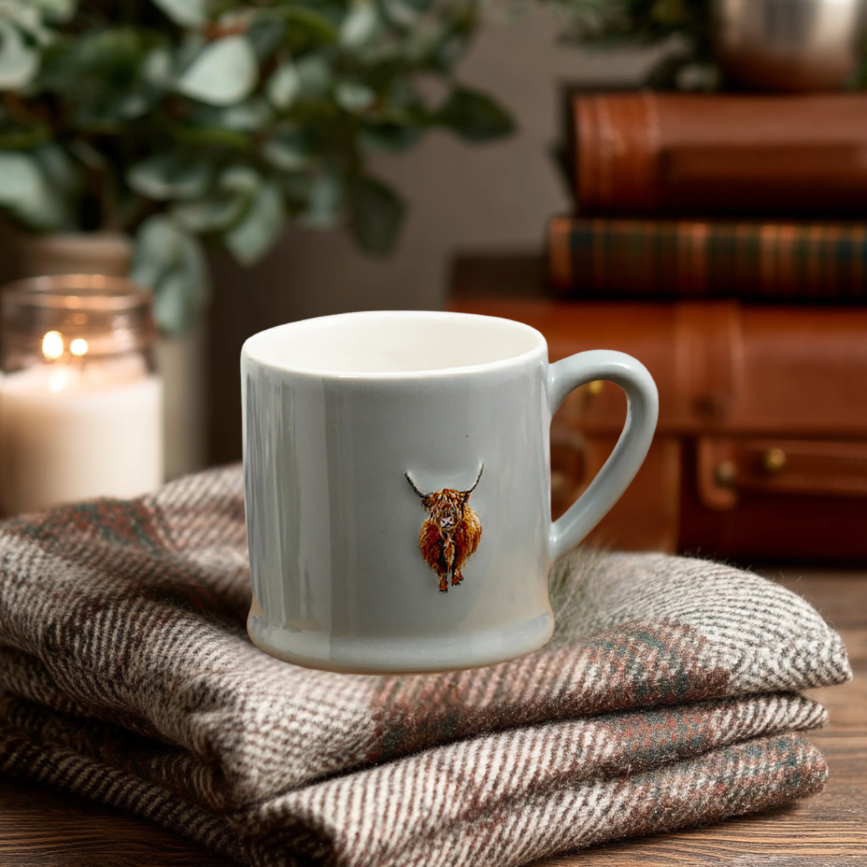 Highland Cow mug on a woven fabric surface with books and a candle in the background.