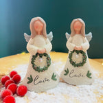 Two ceramic Christmas angel figurines, one holding a green wreath with 'Love' written on it, and the other holding a white wreath with 'C' written on it, standing on a bed of fake snowflakes.