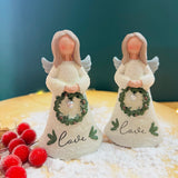 Two ceramic Christmas angel figurines, one holding a green wreath with 'Love' written on it, and the other holding a white wreath with 'C' written on it, standing on a bed of fake snowflakes.