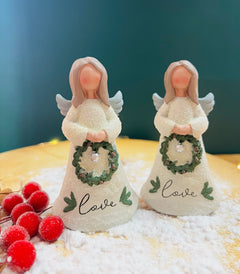 Two ceramic Christmas angel figurines, one holding a green wreath with 'Love' written on it, and the other holding a white wreath with 'C' written on it, standing on a bed of fake snowflakes.