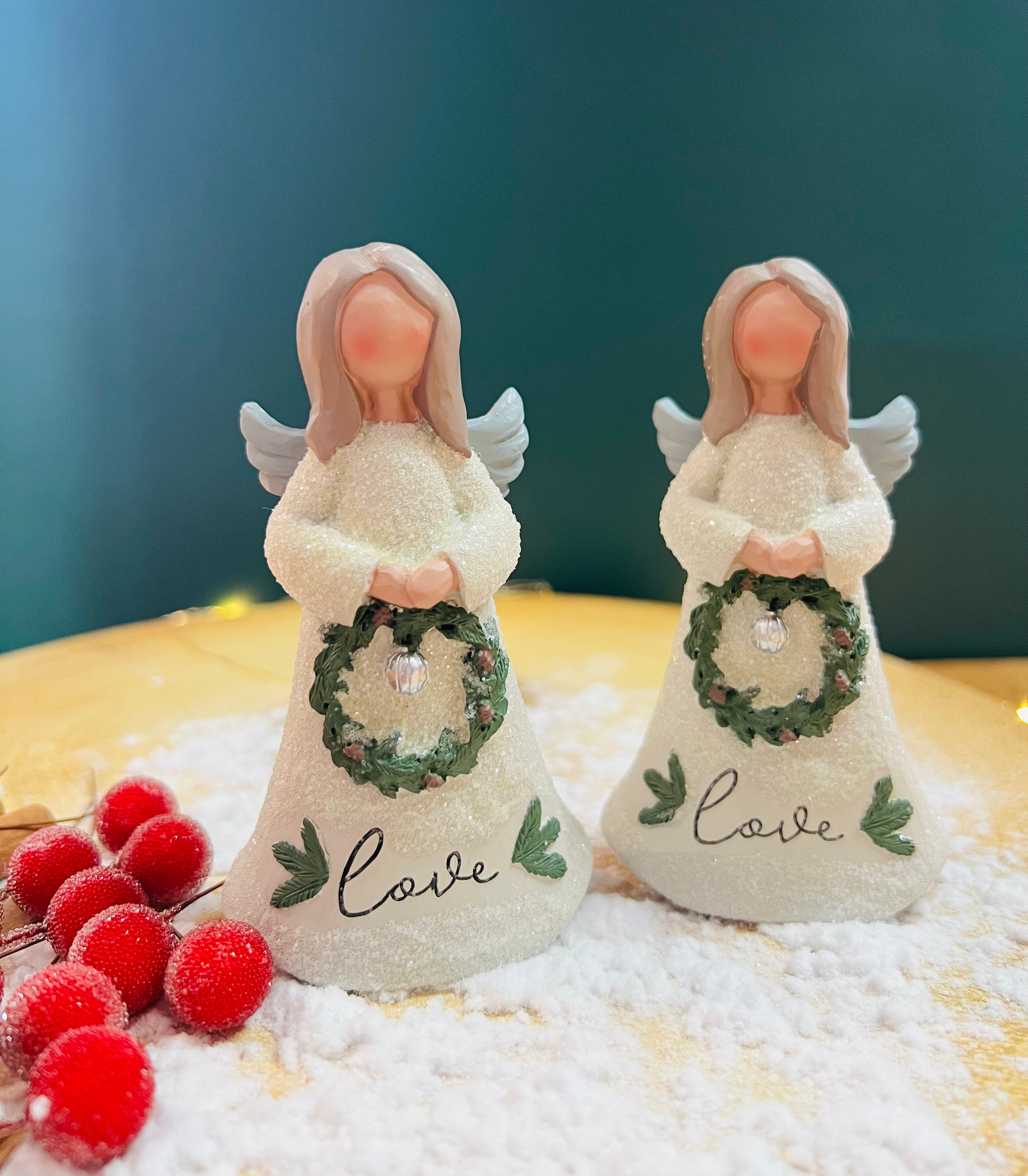 Two ceramic Christmas angel figurines, one holding a green wreath with 'Love' written on it, and the other holding a white wreath with 'C' written on it, standing on a bed of fake snowflakes.