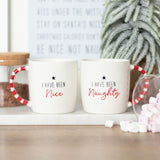 Two white ceramic mugs with red striped handles, one labeled 'I HAVE BEEN NAUGHTY' and the other 'I HAVE BEEN NICE'. The mugs are placed on a white surface with a decorative plant and candy canes in the background.