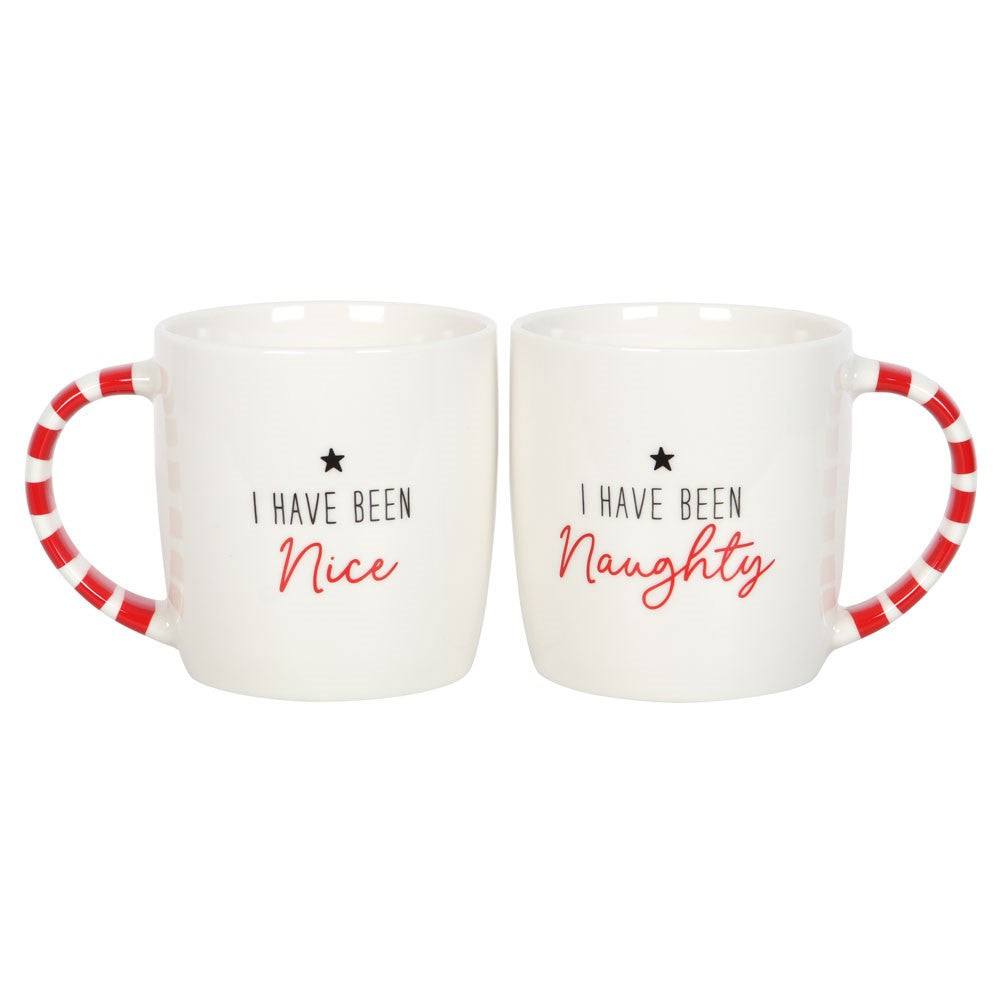 Two white ceramic mugs with red striped handles, one labeled 'I HAVE BEEN NAUGHTY' and the other 'I HAVE BEEN NICE'. The mugs are placed on a white surface with a decorative plant and candy canes in the background.