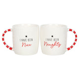 Two white ceramic mugs with red striped handles, one labeled 'I HAVE BEEN NAUGHTY' and the other 'I HAVE BEEN NICE'. The mugs are placed on a white surface with a decorative plant and candy canes in the background.