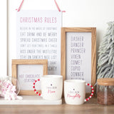 Two white ceramic mugs with red striped handles, one labeled 'I HAVE BEEN NAUGHTY' and the other 'I HAVE BEEN NICE'. The mugs are placed on a white surface with a decorative plant and candy canes in the background.