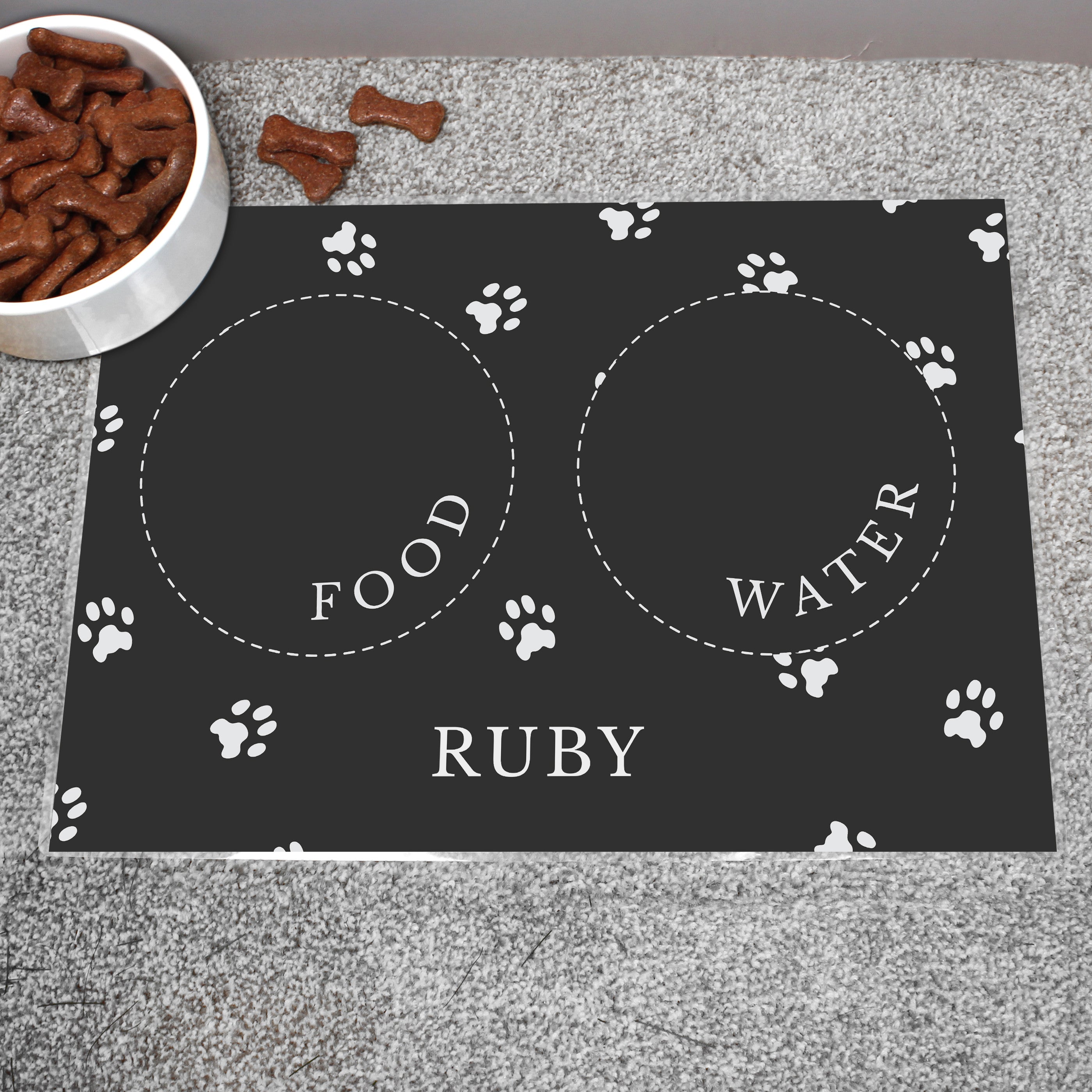 Black personalised pet bowl mat, with a bowl full of dog treats to the side 