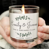A personalised jar candle with a botanical design, customised with the names 'Emily & James' and the phrase 'This is our happy place'. The candle is being held in hands and is lit, showing the flame.