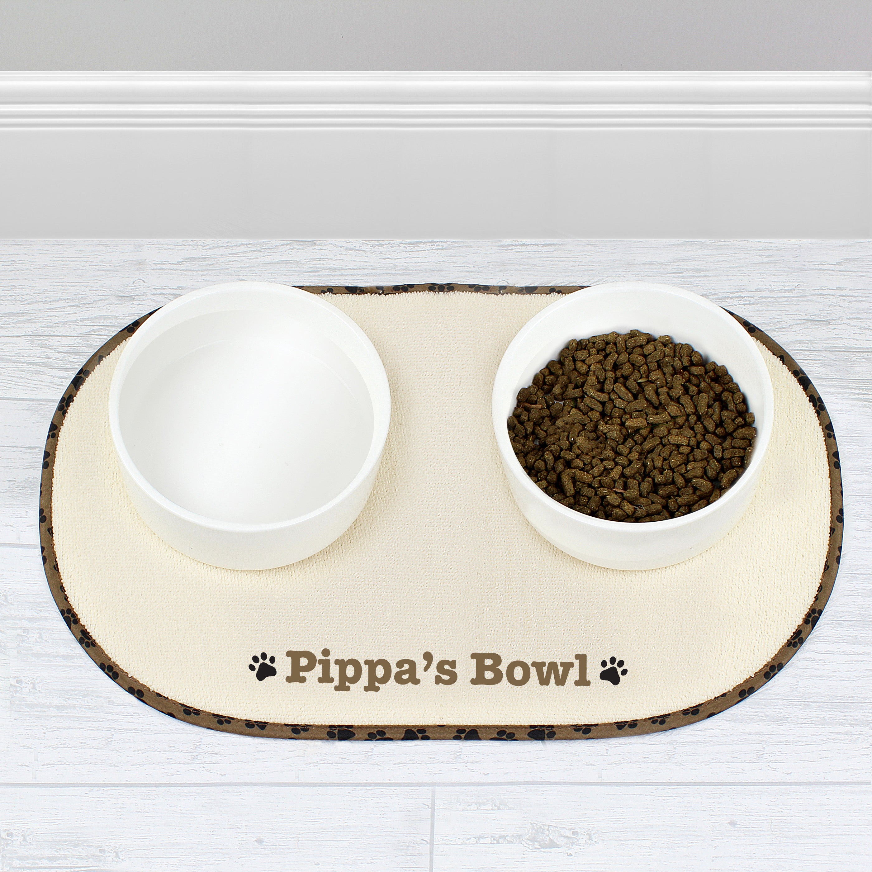 A beige microfibre pet bowl mat with a brown paw print design, personalised with the name 'Pippa', alongside two white bowls filled with pet food.
