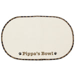 A beige microfibre pet bowl mat with a brown paw print design, personalised with the name 'Pippa', alongside two white bowls filled with pet food.