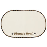 A beige microfibre pet bowl mat with a brown paw print design, personalised with the name 'Pippa', alongside two white bowls filled with pet food.