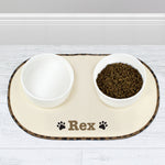 A beige microfibre pet bowl mat with a brown paw print design, personalised with the name 'Pippa', alongside two white bowls filled with pet food.