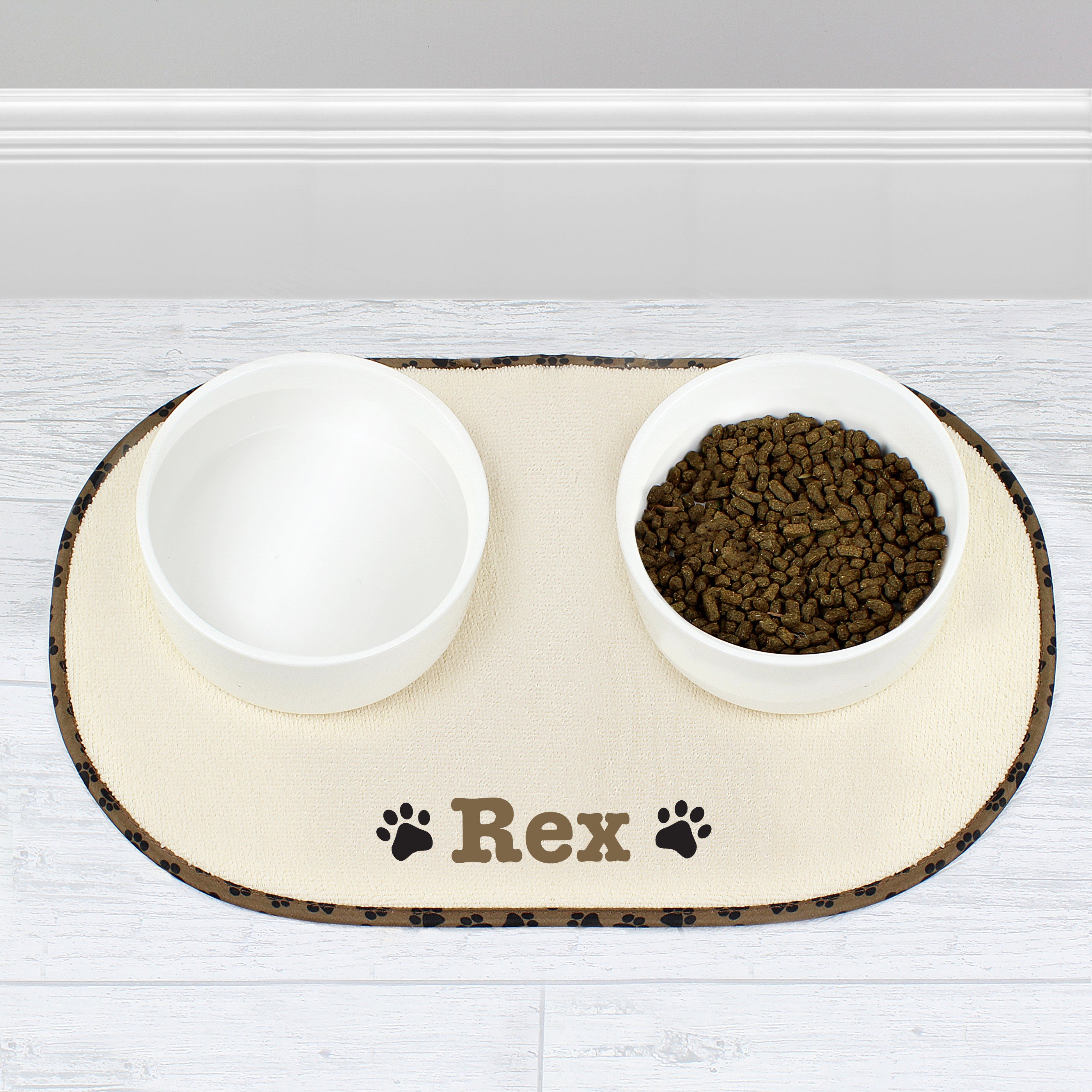 A beige microfibre pet bowl mat with a brown paw print design, personalised with the name 'Pippa', alongside two white bowls filled with pet food.
