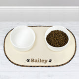 A beige microfibre pet bowl mat with a brown paw print design, personalised with the name 'Pippa', alongside two white bowls filled with pet food.