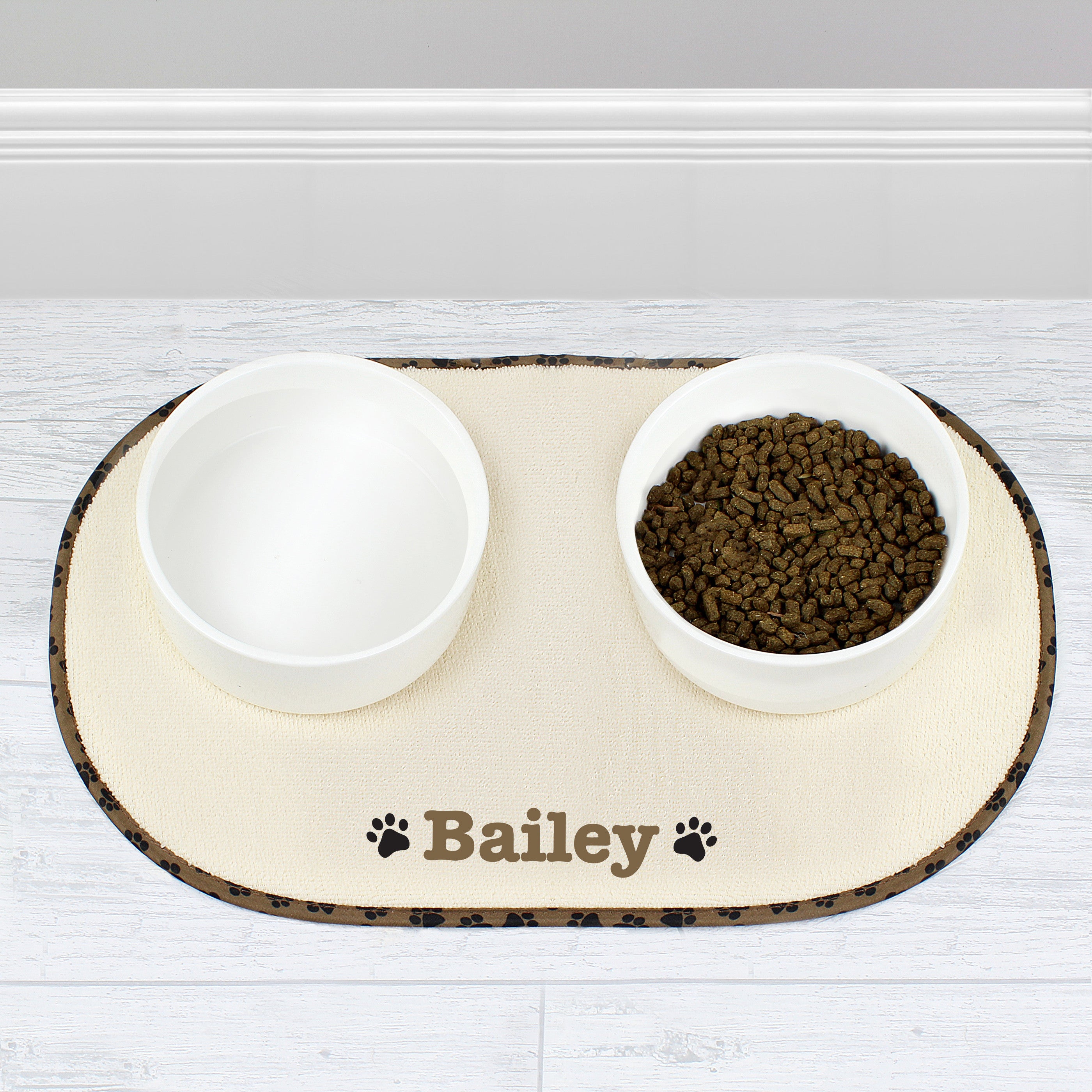 A beige microfibre pet bowl mat with a brown paw print design, personalised with the name 'Pippa', alongside two white bowls filled with pet food.