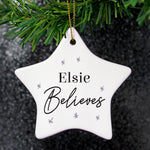 A white ceramic star-shaped decoration with the word 'ELSIE BELIEVES' printed in black, hanging from a golden ribbon on a green pine branch.
