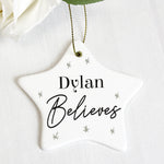 A white ceramic star-shaped decoration with the word 'ELSIE BELIEVES' printed in black, hanging from a golden ribbon on a green pine branch. 01