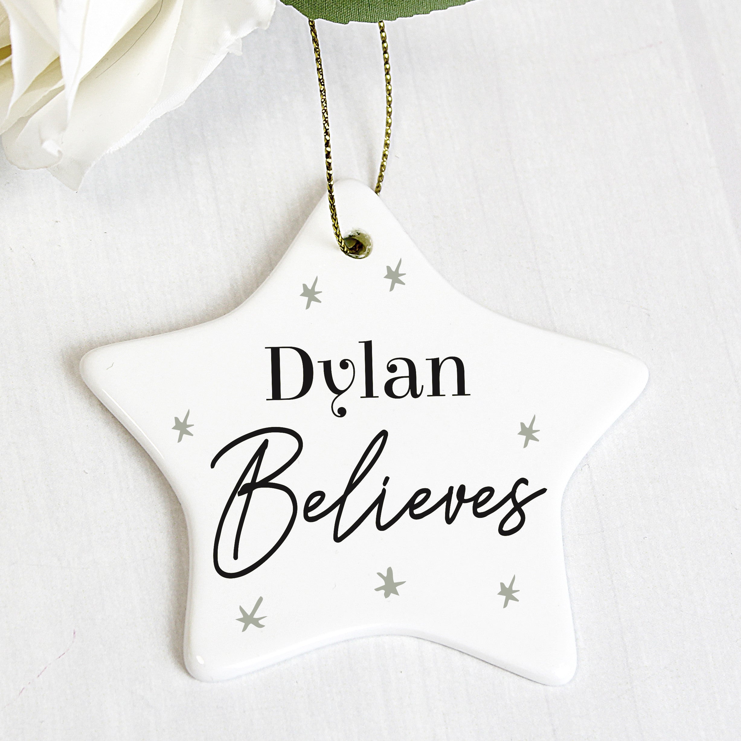 A white ceramic star-shaped decoration with the word 'ELSIE BELIEVES' printed in black, hanging from a golden ribbon on a green pine branch. 01