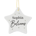 A white ceramic star-shaped decoration with the word 'ELSIE BELIEVES' printed in black, hanging from a golden ribbon on a green pine branch. 02