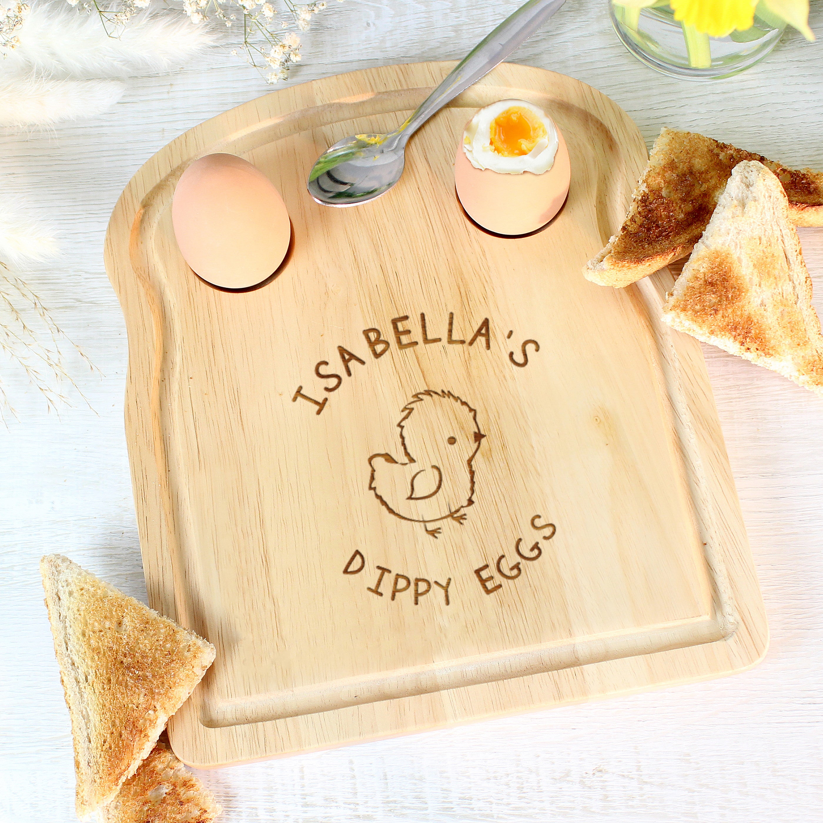 A wooden egg & toast board with a chick design and personalized text 'Isabella's Dippy Eggs' displayed, accompanied by a half-eaten egg and toast on the side.
