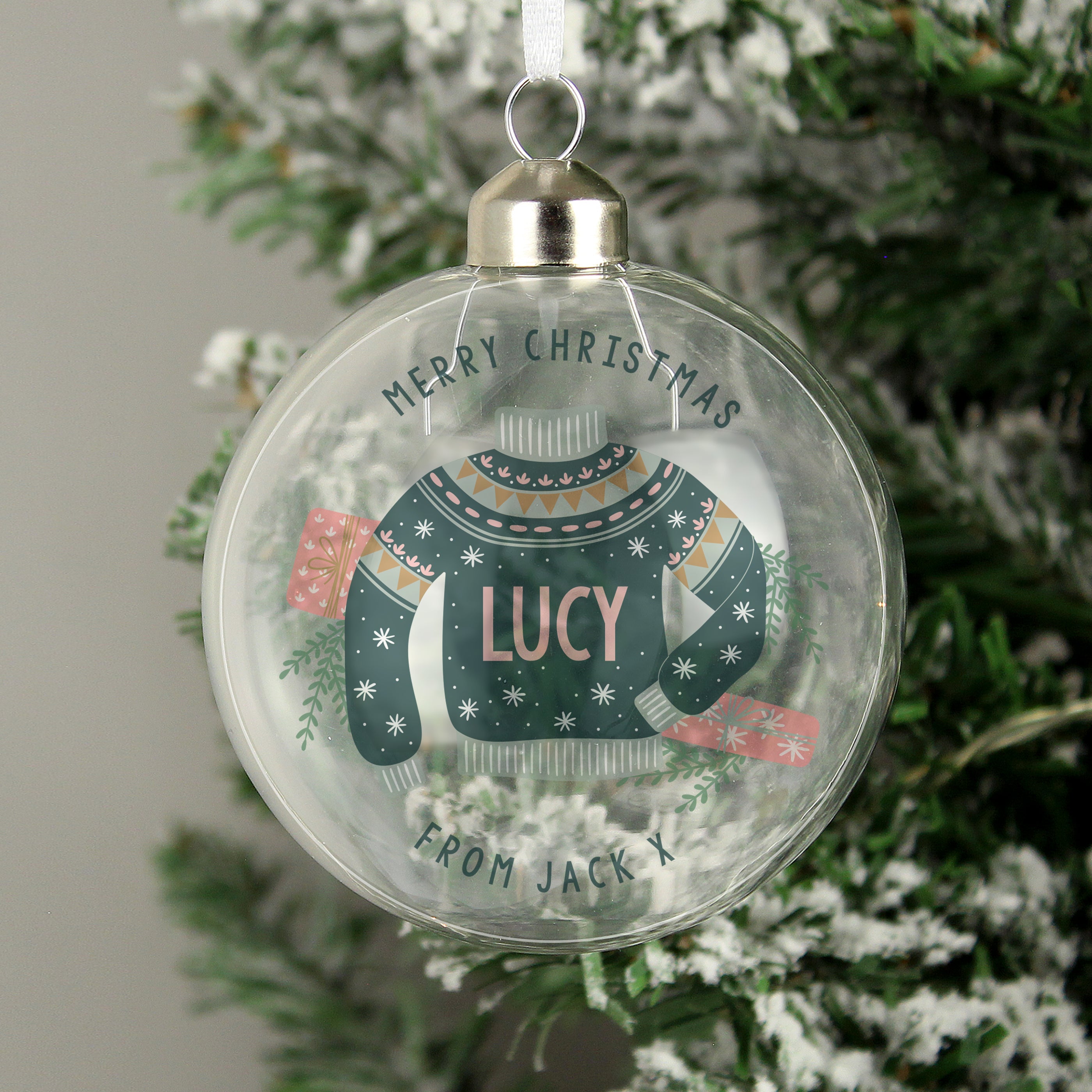 A personalised glass bauble with a Christmas tree and a green sweater design with 'Merry Christmas' and a name 'LUCY' printed on it, hanging from a tree with snow on the ground.