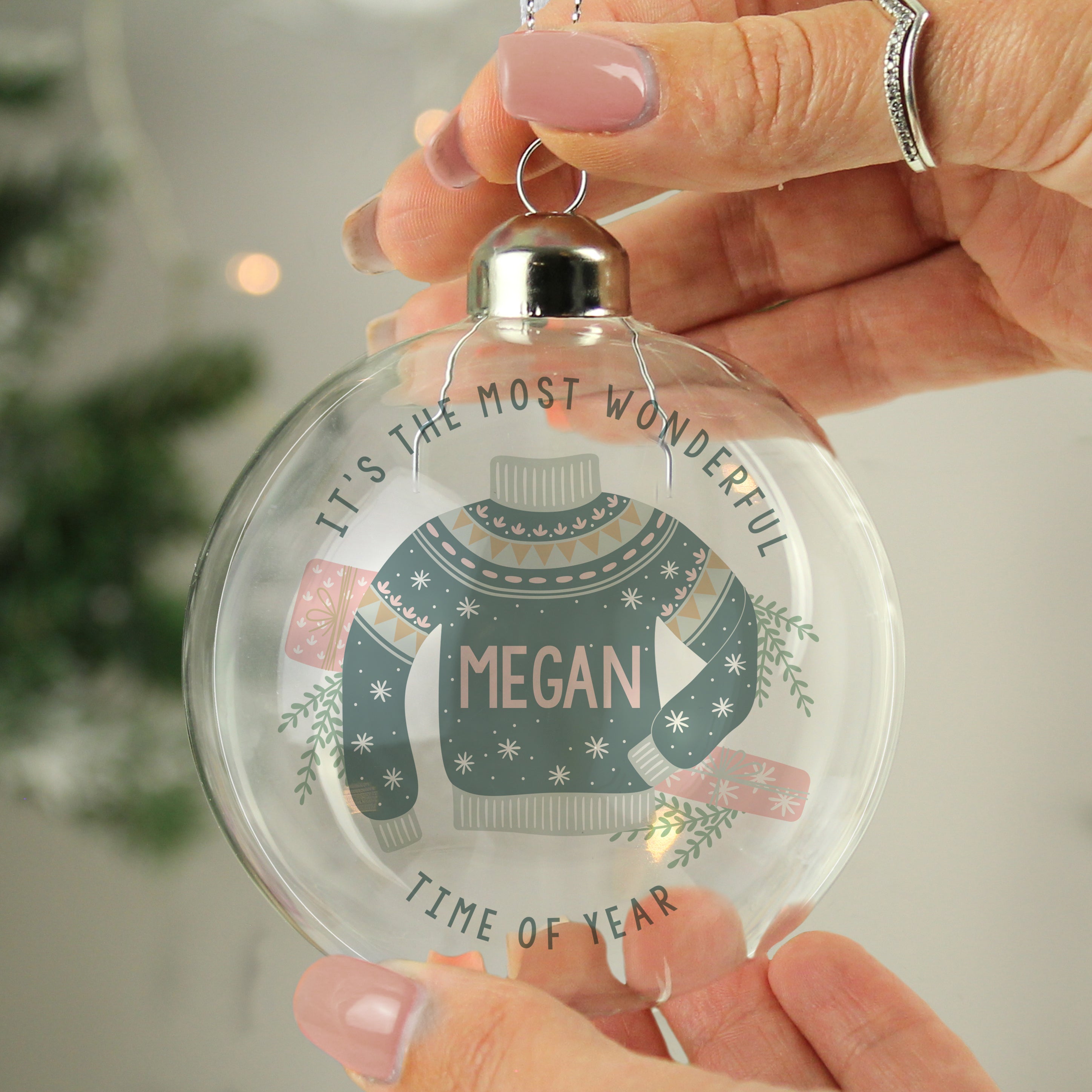 A personalised glass bauble with a Christmas tree and a green sweater design with 'Merry Christmas' and a name 'LUCY' printed on it, hanging from a tree with snow on the ground. 01