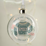 A personalised glass bauble with a Christmas tree and a green sweater design with 'Merry Christmas' and a name 'LUCY' printed on it, hanging from a tree with snow on the ground. 03