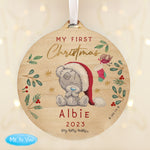 A round wooden decoration featuring an illustration of a teddy bear with a Santa hat, surrounded by Christmas-themed elements like stars, berries, and the text 'My First Christmas'.