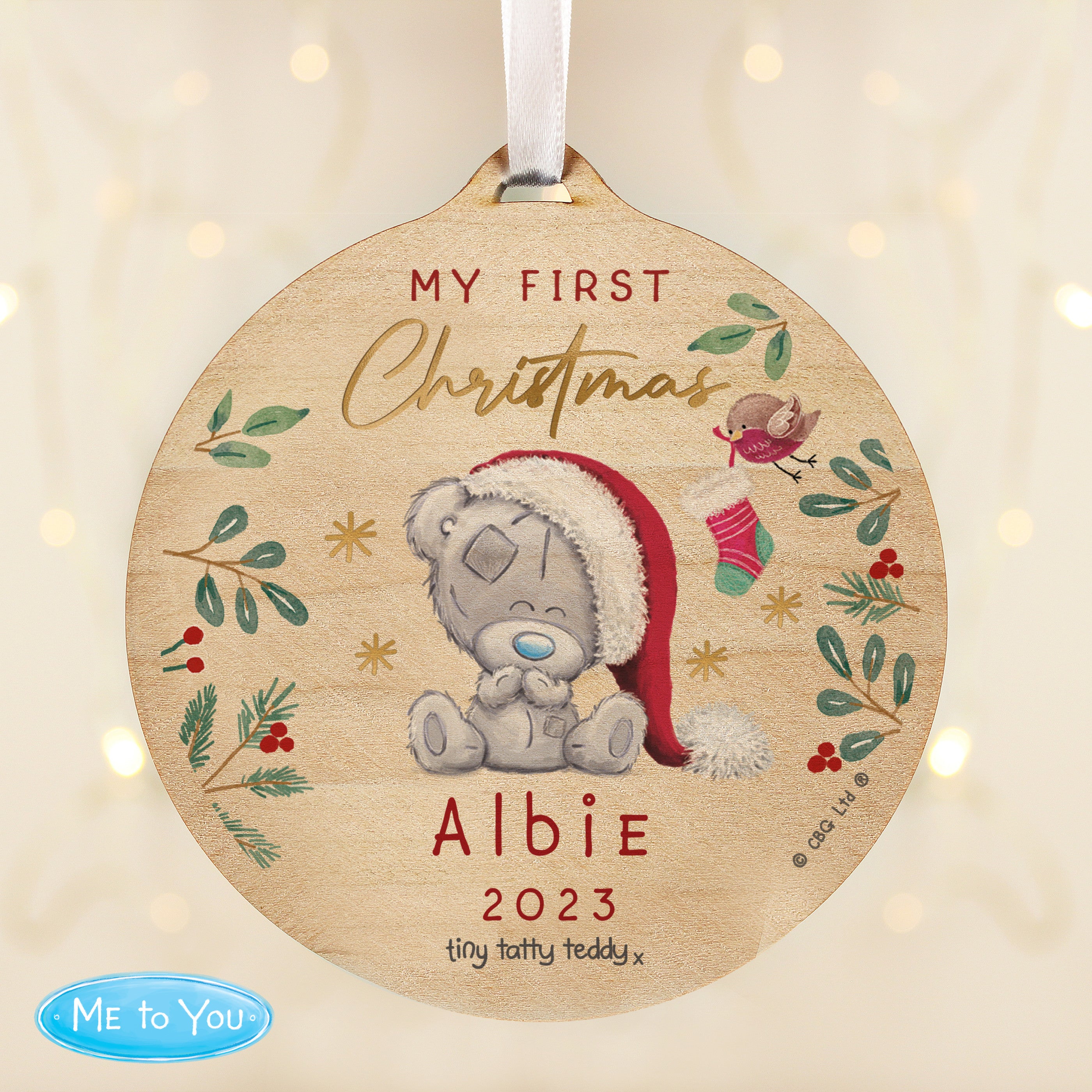 A round wooden decoration featuring an illustration of a teddy bear with a Santa hat, surrounded by Christmas-themed elements like stars, berries, and the text 'My First Christmas'.