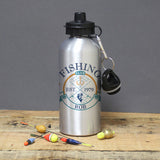 A silver aluminium drinks bottle with black lid and custom text 'FISHING CLUB EST. 1979 ROB' printed on it, displayed alongside fishing accessories on a wooden surface.