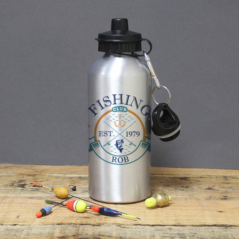 A silver aluminium drinks bottle with black lid and custom text 'FISHING CLUB EST. 1979 ROB' printed on it, displayed alongside fishing accessories on a wooden surface.