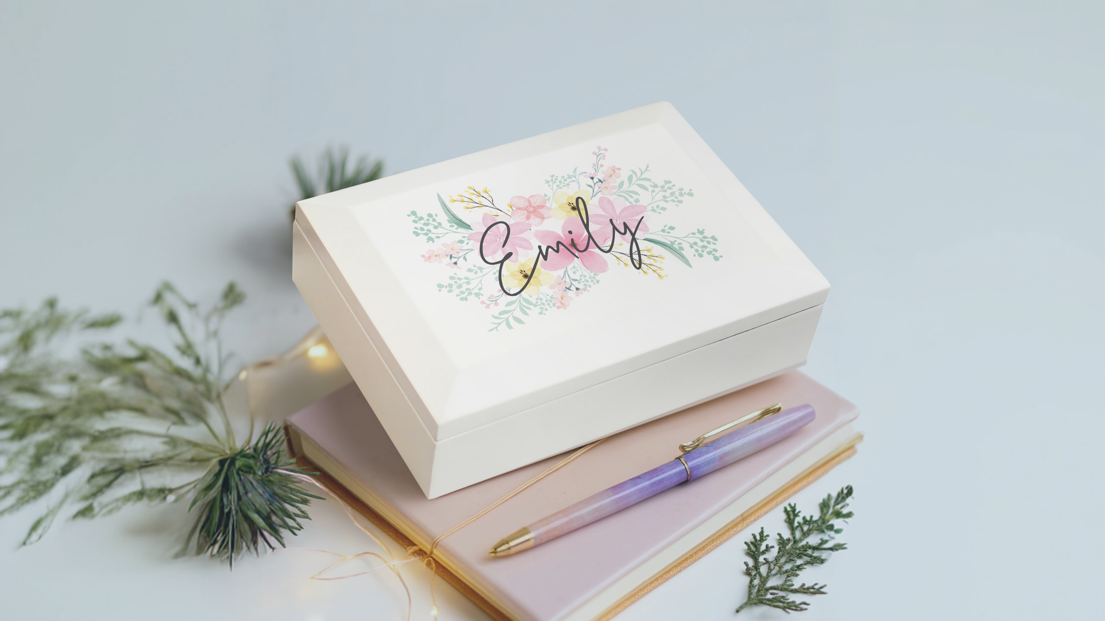 Personalised Floral Wooden Jewellery Box  and 'Emily' text, placed on a notebook with a pen, on a light blue background.