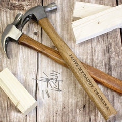 A personalised hammer with a wooden handle and metal head, engraved with the text 'Daddy', along with a few nails and pieces of wood scattered around it.